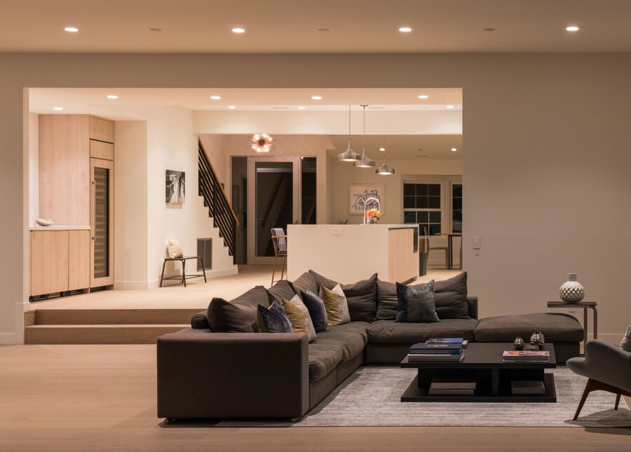 A living room overseeing a luxurious kitchen. There are different color temperatures in the lighting of each room. 