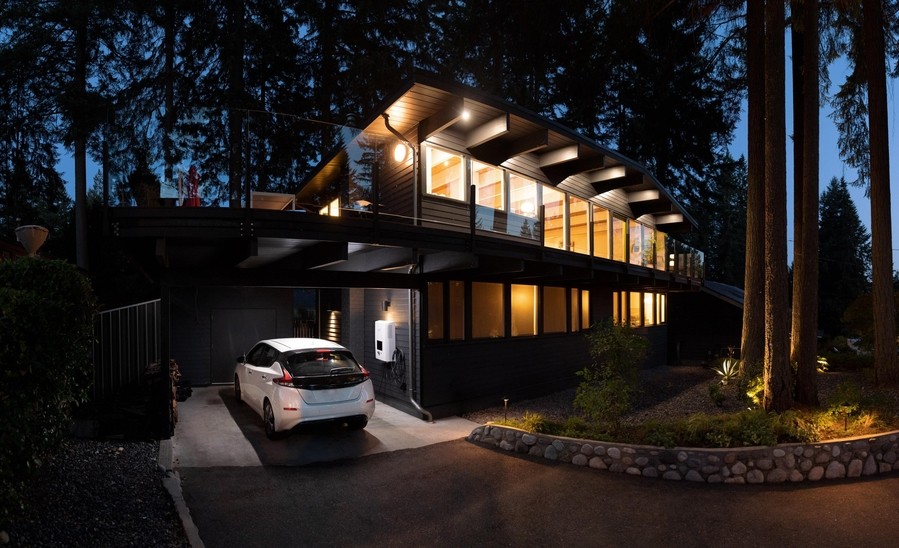Modern home at night illuminated by warm lighting, surrounded by woods. An electric car is parked in the driveway. 